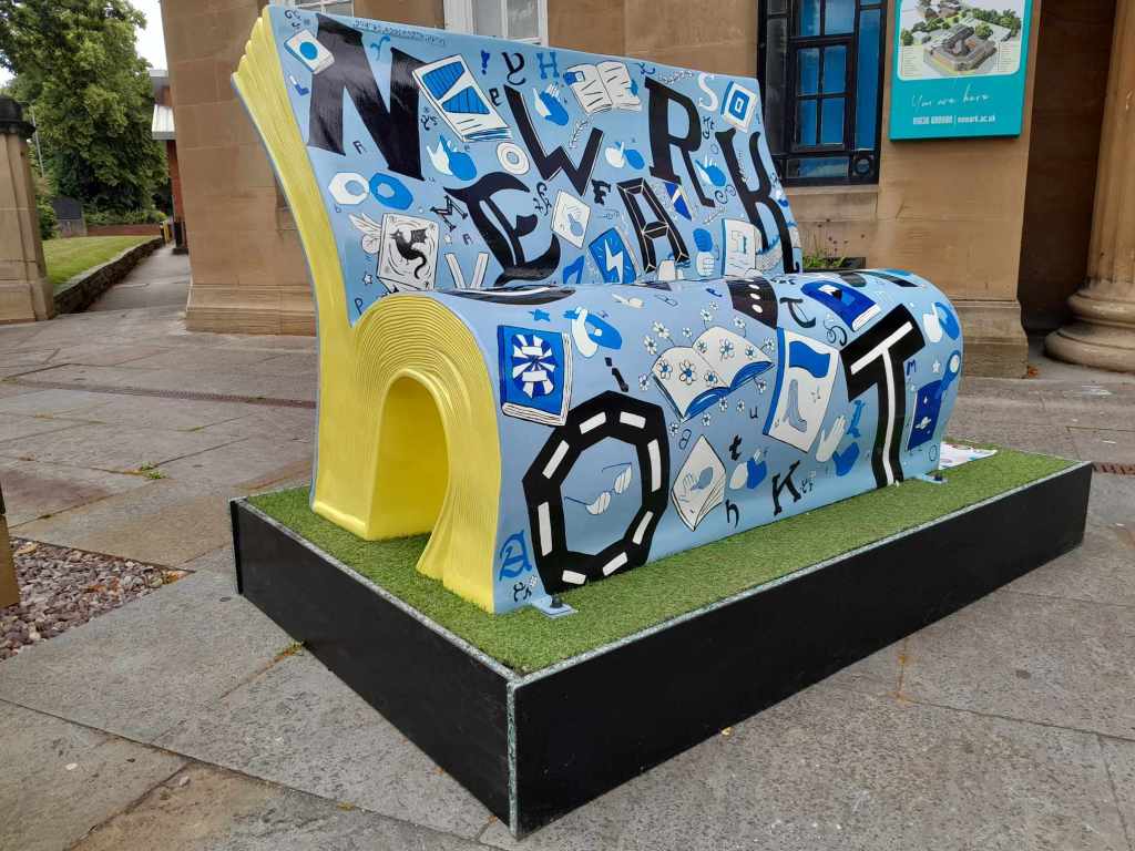 A front and side view of the Alphabets Bookbench by Charlotte Adderley.

The sides are a bright yellow and the front is light blue with black and dark blue lettering. All hand painted alongside illustrations of BSL fingerspelling the alphabet and UEB. The large black lettering says Newark and there are words in grade 1 UEB along the top that read Newark Bookbench Trail. 
