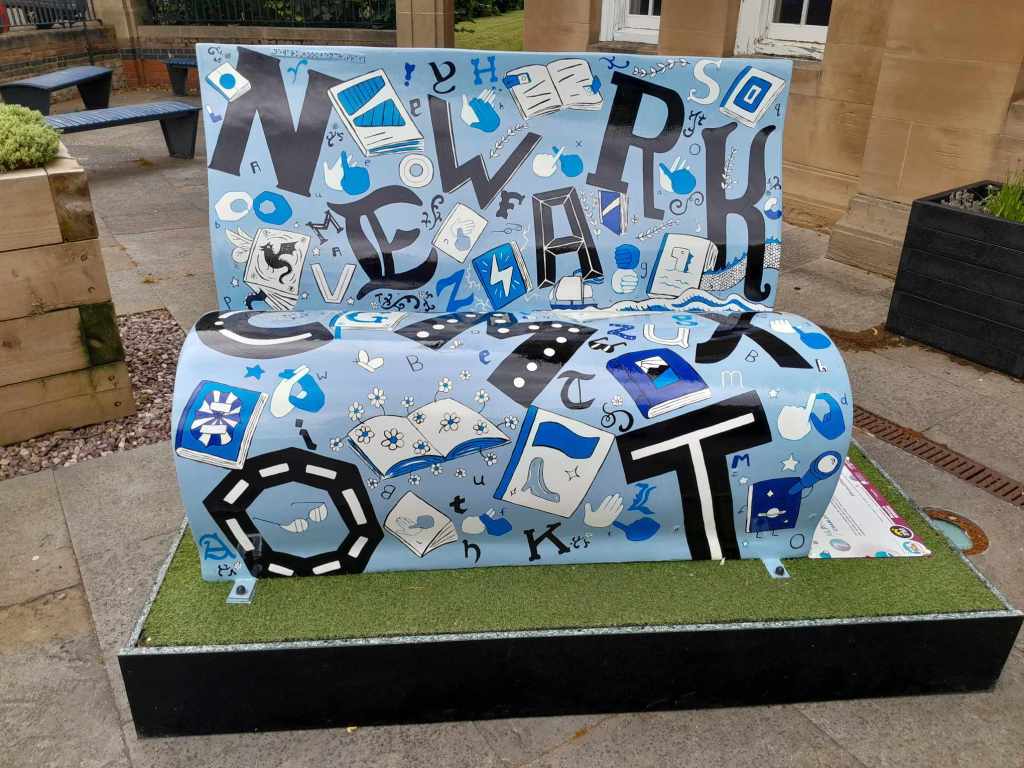 A front view of the Alphabets book bench by Charlotte Adderley. The bench is light blue with black and dark blue lettering in different font styles hand painted alongside illustrations of British Sign Language Fingerspelling alphabet. There are doodles of books around the alphabets of sea creatures fairytale books, flowers growing out of book pages and a ship sailing across the waves of a story. The bench also features the UEB alphabet (mostly on the back of the bench). The large lettering on the front of the bench spells out Newark and along the top of the front of the bench is the words: Newark Bookbench Trail in UEB grade 1. 
