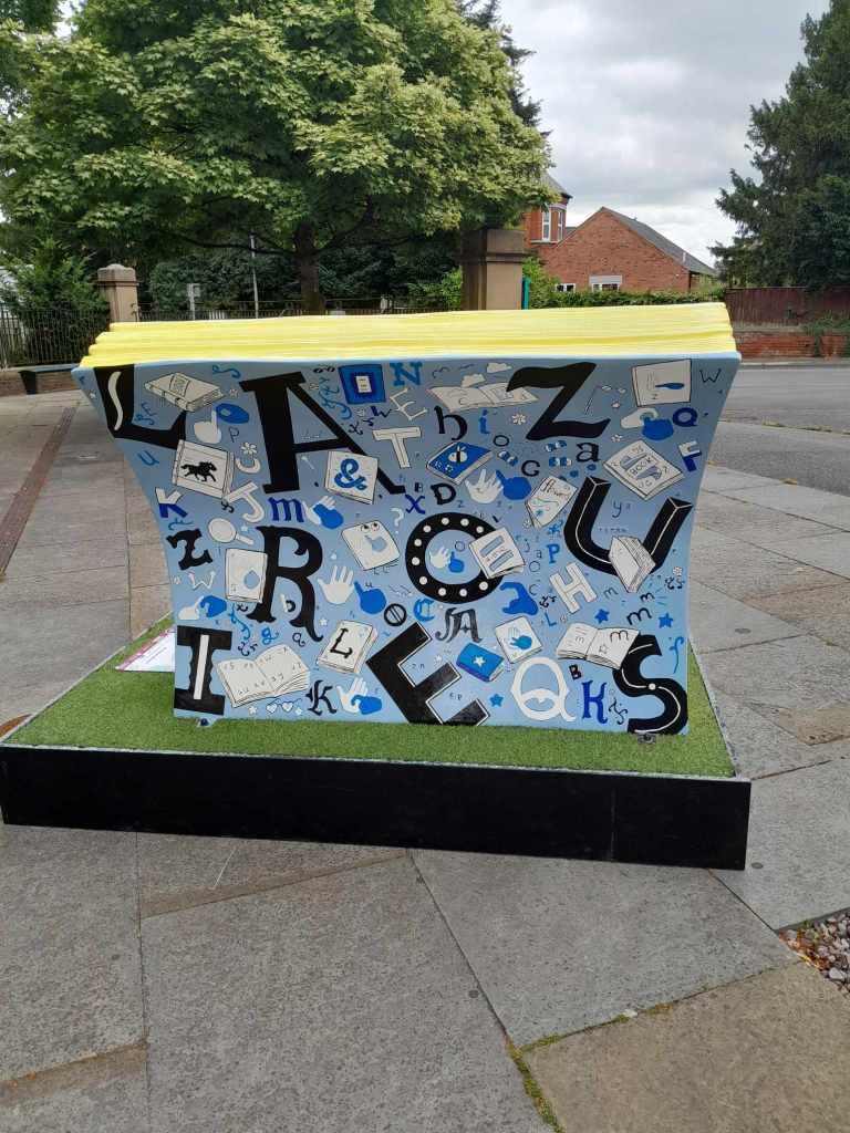 A back view of the Alphabets book bench by Charlotte Adderley. The bench is light blue with black and dark blue lettering in different font styles hand painted alongside illustrations of British Sign Language Fingerspelling alphabet. There are doodles of books around the alphabets of sea creatures fairytale books, flowers growing out of book pages and a ship sailing across the waves of a story. The bench also features the UEB alphabet (mostly on the back of the bench). The large lettering on the front of the bench spells out Newark and along the top of the front of the bench is the words: Newark Bookbench Trail in UEB grade 1. 