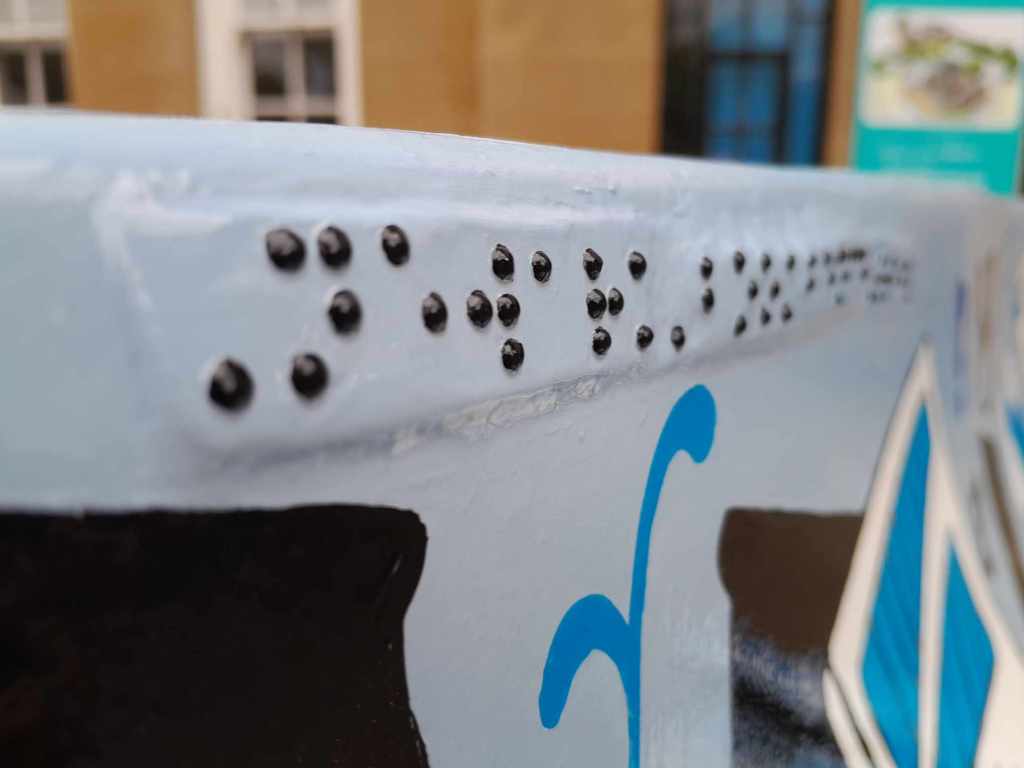 A close up photograph of the braille on the front of the bench that says Newark Bookbench Trail in grade 1 Unified English Braille.